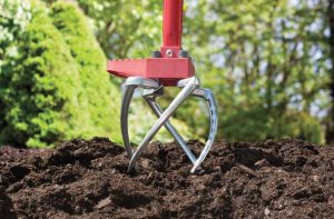 A red-handled manual garden cultivator with metal tines is digging into dark soil in a garden, ideal for package design or Advertising shoots, with green foliage and trees softly blurred in the background.