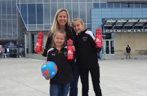 A woman and two girls stand smiling outside a modern stadium entrance. The girls hold red water bottles with eye-catching packaging and wear black sports jackets; one also holds a blue soccer ball with red and white patterns.