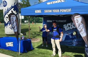 Two people in blue Sport Shake shirts stand at a promotional tent with marketing banners and a branded table outdoors. One waves while the other smiles and gives a thumbs up. The tent reads, SHOW YOUR POWER!.