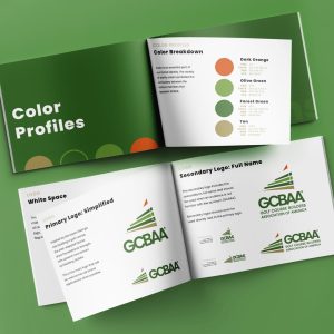 Two open booklets on a green surface display branding guidelines, including color profiles and logo variations for the Golf Course Builders Association of America (GCBAA), with green-themed designs and text tailored for advertising and packaging applications.