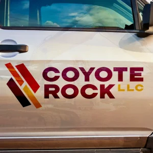 The image features a vehicle door with a bold branding logo reading Coyote Rock LLC. The design incorporates abstract stone shapes in red, orange, and purple. The doors reflection reveals construction equipment, enhancing the industrial vibe.